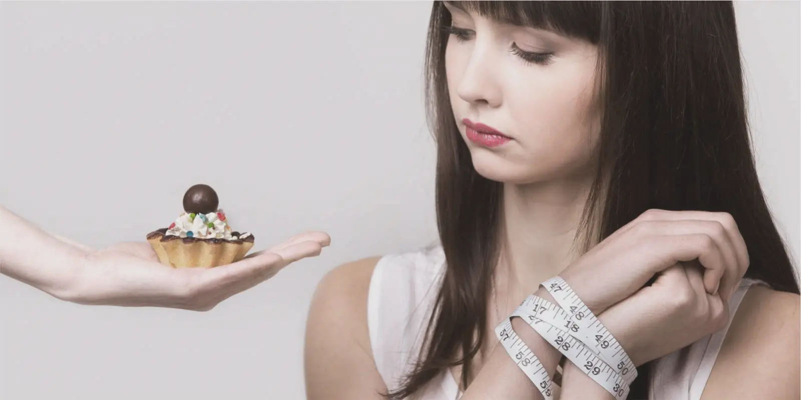 Uma mulher com as mãos amarradas com uma fita métrica. Ela quer comer o doce que está sendo oferecido. Dieta não é eficaz contra a obesidade.